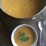 Vegan Red Lentil and Potato Soup in a pot on a table with a bowl of the same soup with cilantro garnish. Background is wooden table. Silver spoon sitting next to the bowl.