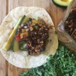 Opened Vegan Burrito sitting on a wooden cutting board next to kale, avocado and extra beans and rice.