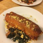 Two plates of Vegan Enchiladas on a white plate with kale on the site and crumbles on top. Plates on a wooden surface.