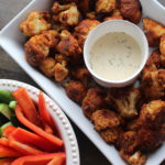 oil-free vegan cauliflower buffalo wings in a white dish with bell peppers and cucumbers in a white dish, and a bowl of ranch dressing.