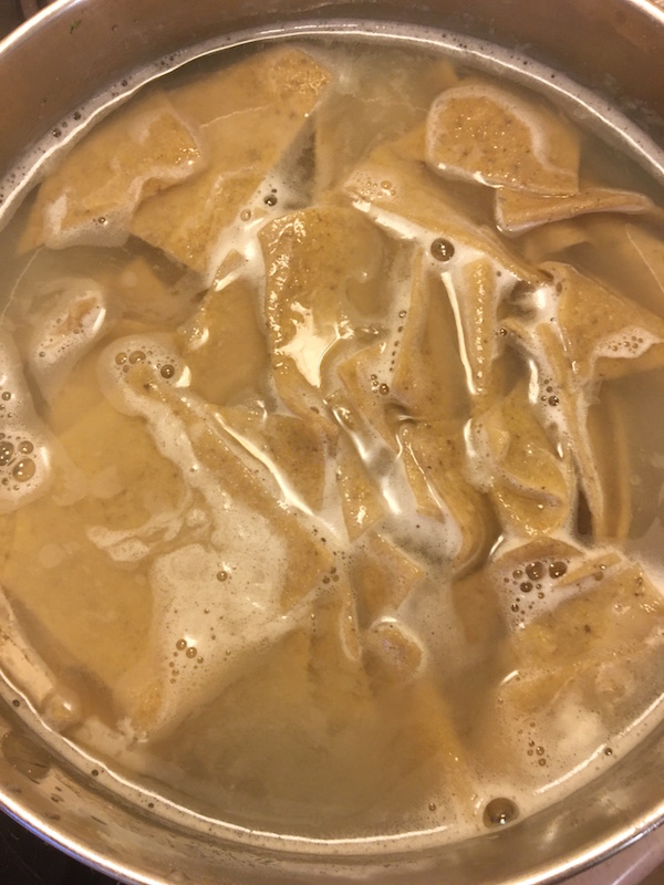 Eggless vegan pasta dough in a large stainless steel pot and water. Foam has risen to the top and pasta appears to be boiling. Pasta looks halfway cooked through. 