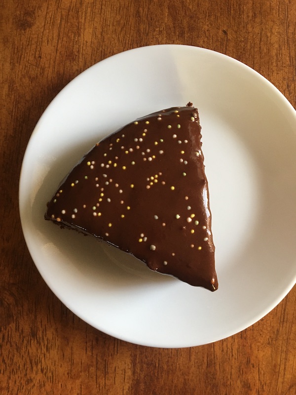 Photo of a delicious piece of vegan chocolate cake made using the recipe below. This cake is oil-free, whole grain as well. Photo of cake shows a piece of cake with chocolate glaze and sprinkles on a small white plate on a dining table. Overhead view. easyveganmealprep.com